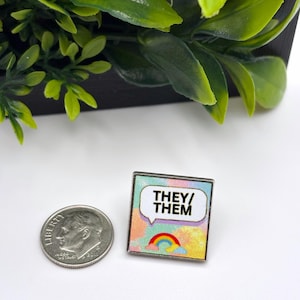 Puede incluir: Un pin cuadrado de esmalte con un fondo de nubes en tonos pastel, un globo de texto blanco con el texto negro THEY/THEM y un pequeño icono de arcoíris debajo, colocado junto a una moneda de diez centavos de EE. UU. para escala.
