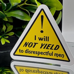 Puede incluir: Una pegatina triangular amarilla con borde negro y un signo de exclamación. El texto dice "I will NOT YIELD to disrespectful men" en fuente negra. La pegatina se refleja en una superficie espejada.