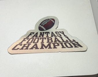 Può includere: Un adesivo con un pallone da football americano e la scritta "FANTASY FOOTBALL CHAMPION" in marrone e bianco. L'adesivo è su un computer portatile bianco.