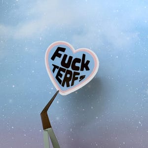 May include: A heart-shaped sticker with the words "Fuck TERFs" in black block letters on a light blue background, outlined in pink. The sticker is held by tweezers against a blue and purple sky background.
