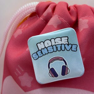 May include: A square, light blue button with the words "NOISE SENSITIVE" in white and blue lettering. Below the text is a graphic of pink and purple headphones. The button is resting on a pink drawstring bag.