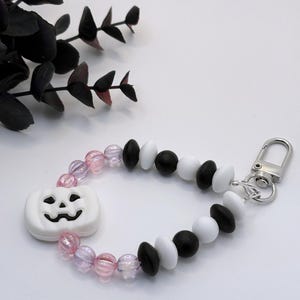 May include: A Halloween-themed keychain featuring a white pumpkin charm with a carved face. The beaded wristlet includes pink, black, and white beads, and a silver-tone clasp. The design is festive and decorative.