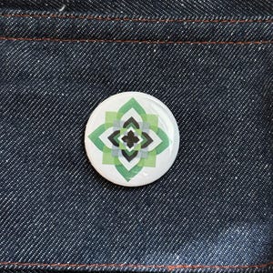 May include: A round button with a white background and a geometric design in shades of green, black, and gray. The button is set against a dark blue denim fabric with orange stitching.