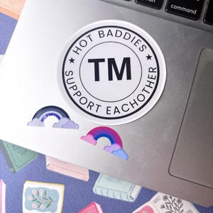 May include: A circular white sticker with a black border and the text HOT BADDIES SUPPORT EACHOTHER surrounding the letters TM in the center. Below the circular sticker are two smaller rainbow-shaped stickers, one in shades of blue and purple.