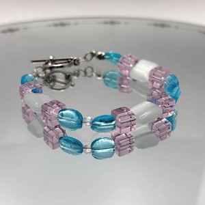 May include: A beaded bracelet featuring a toggle clasp. The bracelet is composed of light blue, lavender, and white beads. The beads are a mix of cube and oval shapes, strung together with silver-colored metal links.