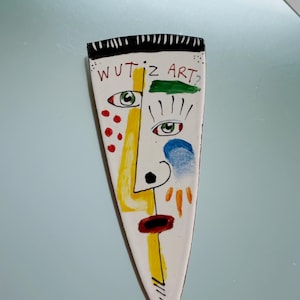 May include: A white ceramic art piece with a painted abstract face. The face features a yellow nose, red lips, and green eyes. The text "WUT'z ART?" is at the top. The piece has a black and white striped top.