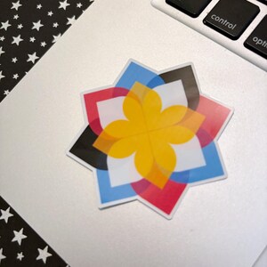 May include: A colorful sticker with a starburst design, featuring overlapping shapes in yellow, red, blue, black, and white. The sticker is placed on a silver laptop, with a black and white star pattern in the background.