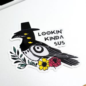 May include: A sticker featuring a black and white bird wearing a black hat, with yellow eyes and accents. The sticker has the text "Lookin' Kinda Sus" and is decorated with flowers and leaves.
