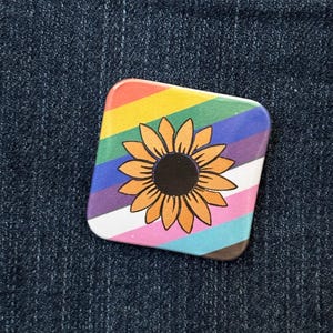 May include: A square pin badge with a sunflower design. The badge has a rainbow background and a sunflower in the centre. The sunflower has orange petals and a black centre. The badge is set against a blue denim background.