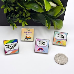 Puede incluir: Cuatro pines cuadrados de esmalte con diseños y textos sobre el orgullo, incluyendo 'Trans rights are human rights', 'Y'all MEANS ALL' y dos pines 'THEY/THEM' con arcoíris. Una moneda de diez centavos de EE. UU. está junto a los pines como referencia.