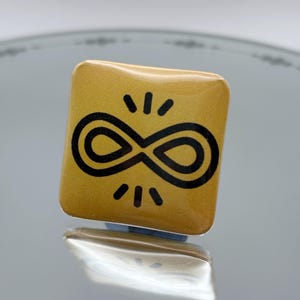 May include: A square, gold-colored pin with a black infinity symbol design. The pin has a glossy finish and is reflected in the surface below. The infinity symbol is surrounded by small, black lines radiating outwards.