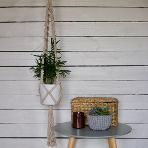 May include: A white ceramic planter with a green plant hanging from a macrame plant hanger. The hanger is made of natural fibre rope and has a long tassel. The planter is sitting on a grey table with a wicker basket and a small potted succulent.