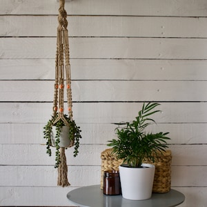 May include: A macrame plant hanger with orange beads and a white pot with a green succulent plant hanging from it. The hanger is against a white wood wall.