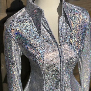 Op de afbeelding: Een zilveren, iriserende blouse met lange mouwen en een ritssluiting. De blouse heeft een subtiel patroon van kleine, iriserende vierkantjes.