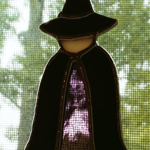 Pode incluir: Um ornamento de vidro colorido com a silhueta de uma bruxa preta com uma túnica de padrão roxo e branco. O ornamento está pendurado em uma janela.
