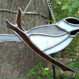 Puede incluir: Atrapasoles de pájaro de vidriera en tonos blanco, gris y negro, posado en una rama marrón. El pájaro está suspendido por una cadena plateada. Una pieza decorativa para una ventana o jardín.
