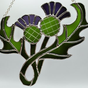 May include: A stained glass window hanging featuring two green and purple thistle flowers with green leaves.