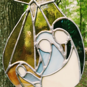 May include: A stained glass ornament depicting the nativity scene with a star, a green and blue robe, and a gold and brown robe.
