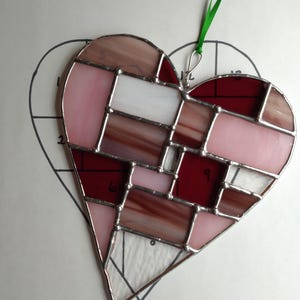 Puede incluir: Un corazón de vidrio coloreado con un diseño de patchwork en tonos de rosa, rojo y marrón. El corazón está colgado de una cinta verde.
