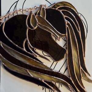 May include: Stained glass artwork depicting a horse's head. The design incorporates brown, amber, and black glass, framed with copper-coloured metal. The piece is suspended by a chain and set against a white background with a pattern.