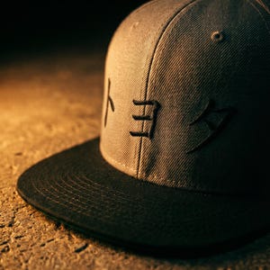 May include: A grey snapback cap with a black peak. The cap has Japanese characters embroidered in black on the front. The hat is made of a textured material and is on a textured surface.