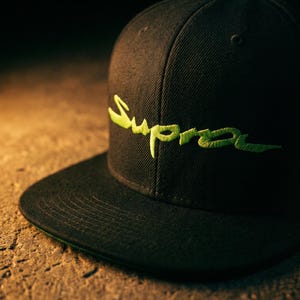 May include: Black baseball cap featuring the word "Supra" embroidered in neon green. The cap has a flat peak and a structured design. The background is a textured, neutral surface.