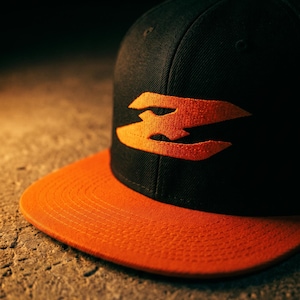 May include: A black baseball cap featuring a flat orange brim and an embroidered orange logo on the front panel. The logo consists of a stylized, jagged horizontal shape. The cap has a button on the top center and a structured crown design.