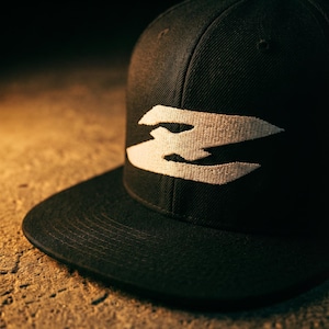 May include: Black baseball cap featuring a white embroidered logo. The cap has a flat peak and a structured crown. The logo is a stylized, abstract design. The cap is made of a textured fabric and rests on a textured surface.