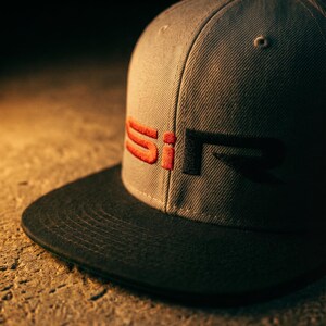 May include: A gray snapback hat with a black brim. The hat features the embroidered letters "SiR" in red and black on the front. The hat is made of a textured fabric and is set against a dark background.