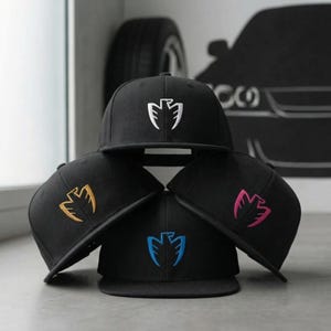 Puede incluir: Cuatro gorras de béisbol negras con visera plana, cada una bordada con un emblema de pájaro estilizado en blanco, dorado, azul y rosa. Las gorras están apiladas sobre una superficie gris claro.