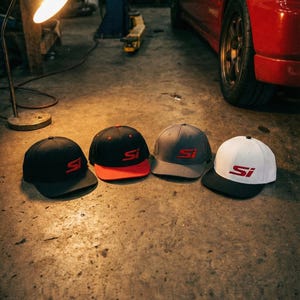 May include: Four baseball caps in a garage setting. The caps are black, red, gray, and white, each with the letters "SI" in red. The caps are on a concrete floor, with a red car in the background.