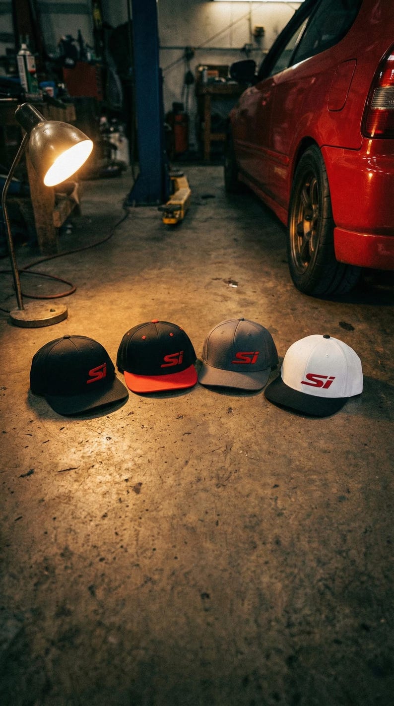 May include: Four baseball caps in a garage setting. The caps are black, red, gray, and white, each with the letters "SI" in red. The caps are on a concrete floor, with a red car in the background.