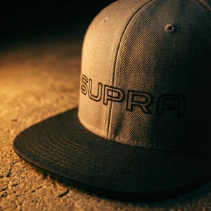May include: A grey baseball cap with a black peak. The word "SUPRA" is embroidered in black on the front. The cap is on a textured, light brown surface.