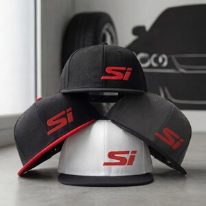 May include: A stack of flat-brimmed baseball caps in black, gray, and white. Each cap features the embroidered letters "Si" in red. One cap has a red brim. The caps are stacked on a light gray surface.