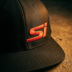 May include: Black baseball cap with a flat brim. The cap features an embroidered red "SI" logo on the front. The hat is made of a textured fabric and sits on a concrete surface.