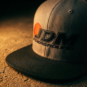 May include: A grey flat-peak cap featuring the embroidered text "JDM UNDERGROUND" in black and orange. The cap has a black peak and a structured fit, suitable for casual wear.