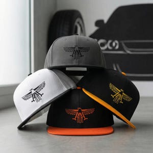 Puede incluir: Un conjunto de gorras de béisbol de visera plana en varios colores: gris, blanco y negro. Cada gorra presenta un diseño de águila bordada. Las gorras negras tienen detalles en naranja y amarillo. Las gorras están sobre una superficie gris claro.