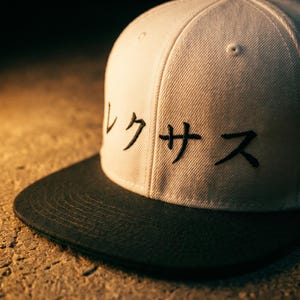 May include: A white baseball cap with a black peak. The cap has Japanese characters embroidered in black on the front. The cap is on a textured surface.
