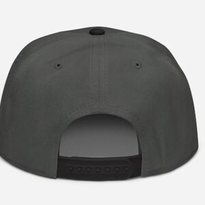 May include: A dark gray baseball cap with a flat brim and a black adjustable snap closure. The cap has a structured crown and a small black button on top. The back of the cap is open.