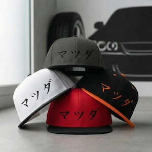 Puede incluir: Una pila de gorras de béisbol de ala plana en varios colores: gris, blanco, negro y rojo. Cada gorra presenta los caracteres japoneses de "Mazda" bordados en la parte delantera. La gorra negra tiene un borde naranja.