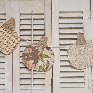 May include: A fall-themed garland featuring three fabric pumpkins. Each pumpkin has a burlap stem and is made from patterned fabric in shades of cream, brown, and floral prints. The pumpkins are strung together with a white string.