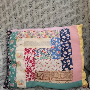 May include: A rectangular throw pillow featuring a patchwork design composed of various fabric scraps in floral, solid, and patterned prints. The colors include shades of pink, blue, green, yellow, white, and brown, arranged in a log cabin style pattern.