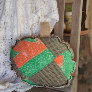 May include: A handmade, quilted pumpkin-shaped pillow with a burlap stem. The pillow features a patchwork design with orange, green, and brown fabrics. The edges are trimmed with a ruffled, cream-colored fabric. The pillow is resting on a wooden chair.
