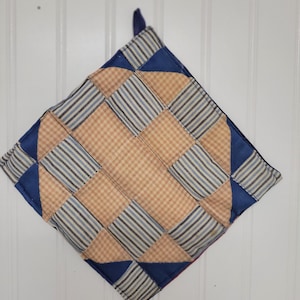 May include: A square quilted potholder featuring a patchwork design of tan and white gingham squares alternating with blue and white striped squares, accented by solid blue triangular corner pieces and a small fabric hanging loop at the top.