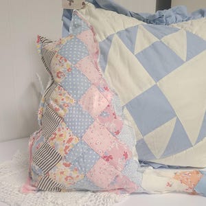 May include: A patchwork cat-shaped pillow in pastel pink, blue, and floral patterns sits next to a blue and white star-patterned pillow. The cat pillow is made of various fabric squares. The pillows rest on a white surface with a lace doily.