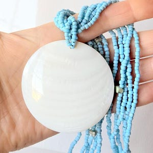 May include: A white, round pendant necklace with a light blue beaded strand. The pendant is large and circular, with a smooth, glossy surface. The necklace is held in a hand against a white background.