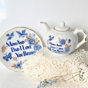 Puede incluir: Un juego de tetera y plato de cerámica blanca con diseños florales y de pájaros azules. El plato y la tetera tienen el texto "Mom and Dad I Love You Both". El juego está sobre un mantel de encaje crema.