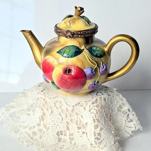 May include: A decorative teapot with a yellow base and gold accents. The teapot features a lid and is adorned with red apples, purple berries, and green leaves. The teapot is resting on a white lace doily.