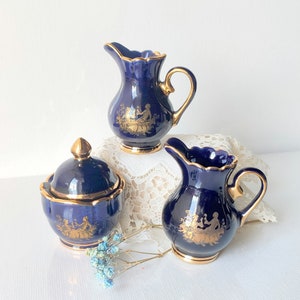May include: A set of three cobalt blue ceramic serving pieces with gold trim. The set includes two small pitchers and a lidded sugar bowl. Each piece is decorated with a gold classical scene.