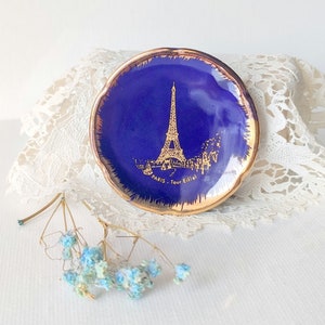 May include: A small, round, cobalt blue dish with a gold rim. The dish features a gold illustration of the Eiffel Tower and the text "PARIS - Tour Eiffel". Decorative item.